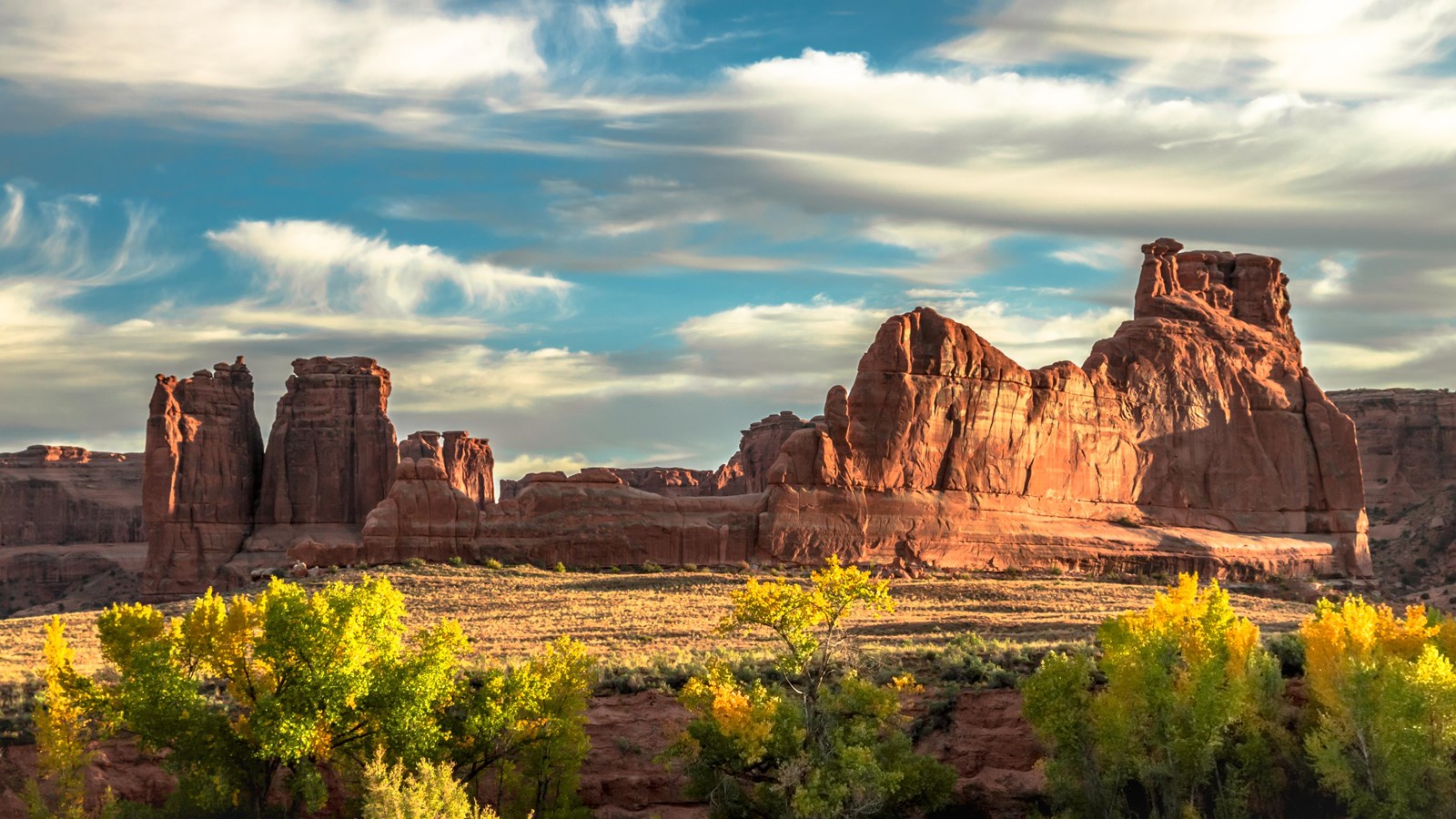 green trees turning yellow beneath red sandstone towers and a cloud-patched sky