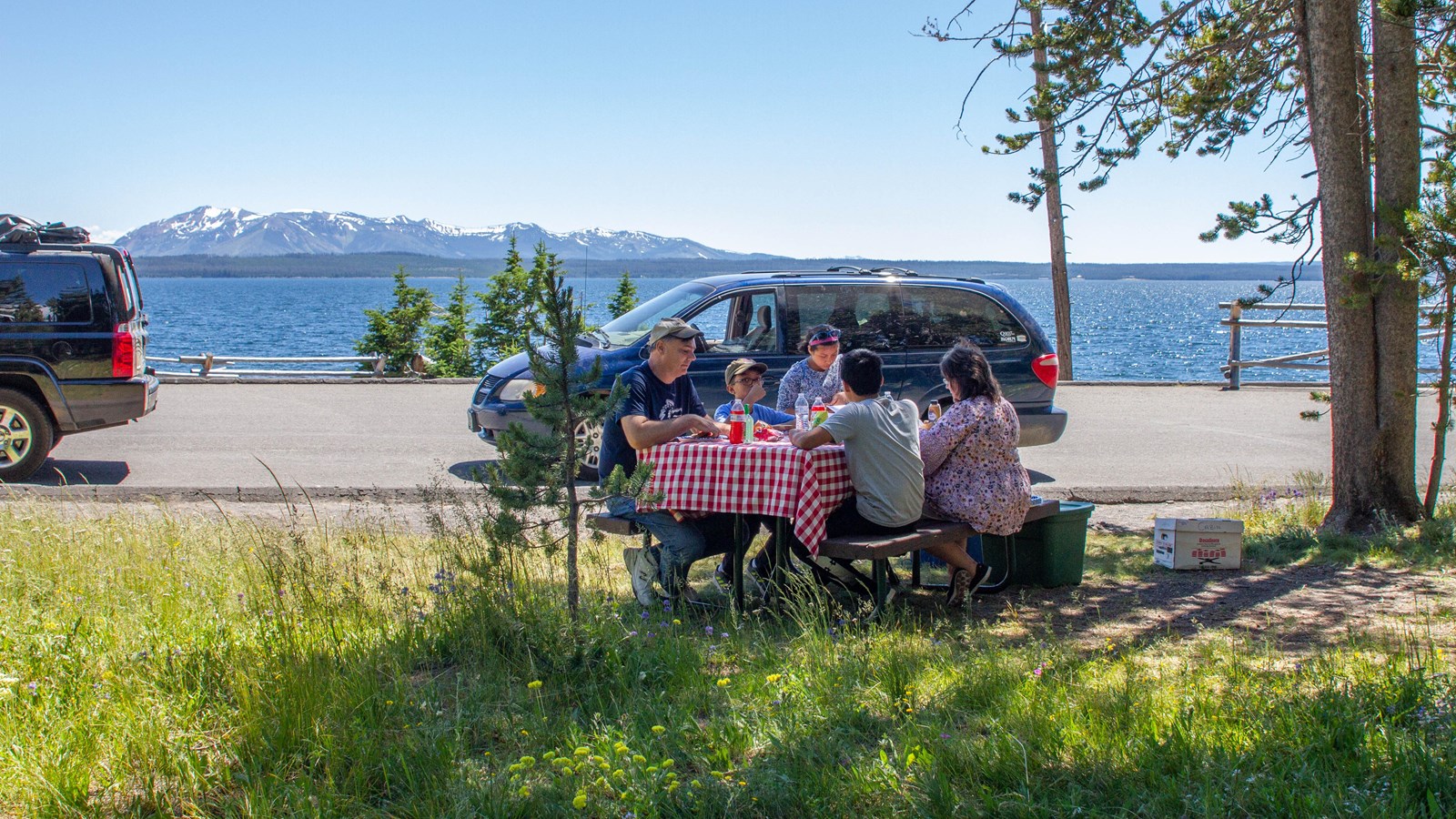 A group of people eating at a picnic table near the shore of a lake