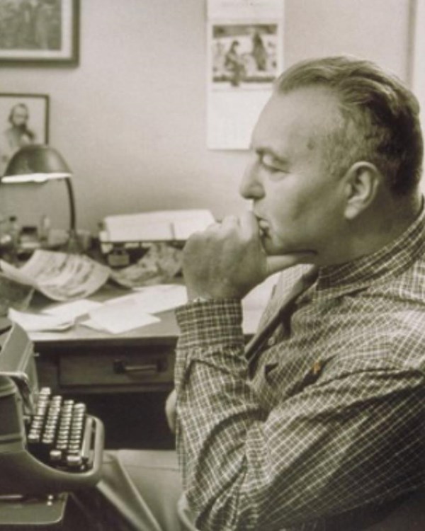 A man is seated in at a desk with a typewriter; his chin is resting on his hand in a thoughtful pose