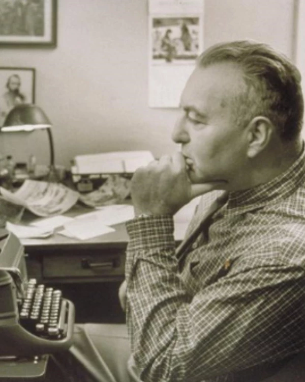 A man is seated in at a desk with a typewriter; his chin is resting on his hand in a thoughtful pose