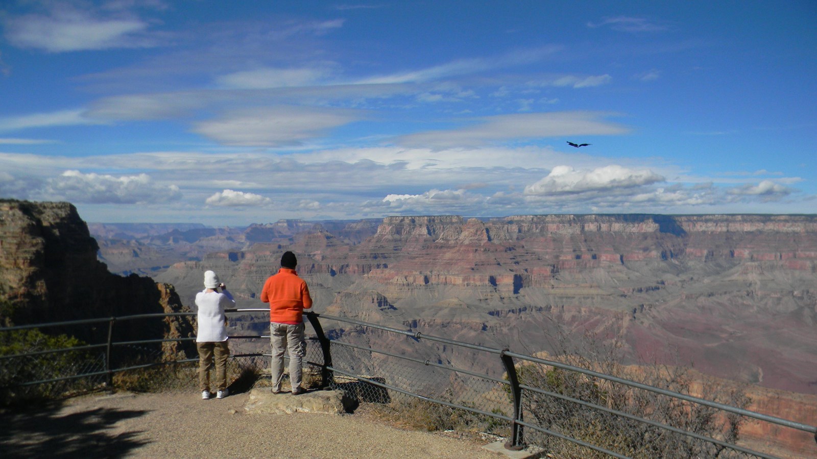 Two people stand at a metal railing admiring a view of Grand Canyon under a blue sky.