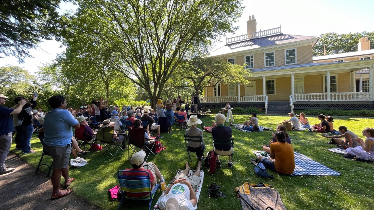 Outdoor crowd listens to music on a shaded lawn