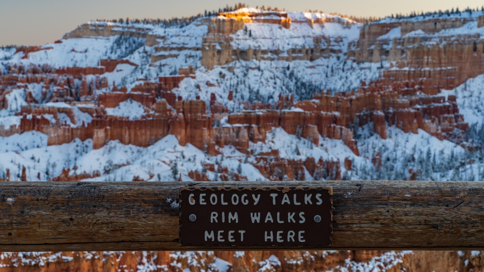 A wooden routed sign reads Geology Talks Rim Walks Meet Here with a snowy landscape beyond