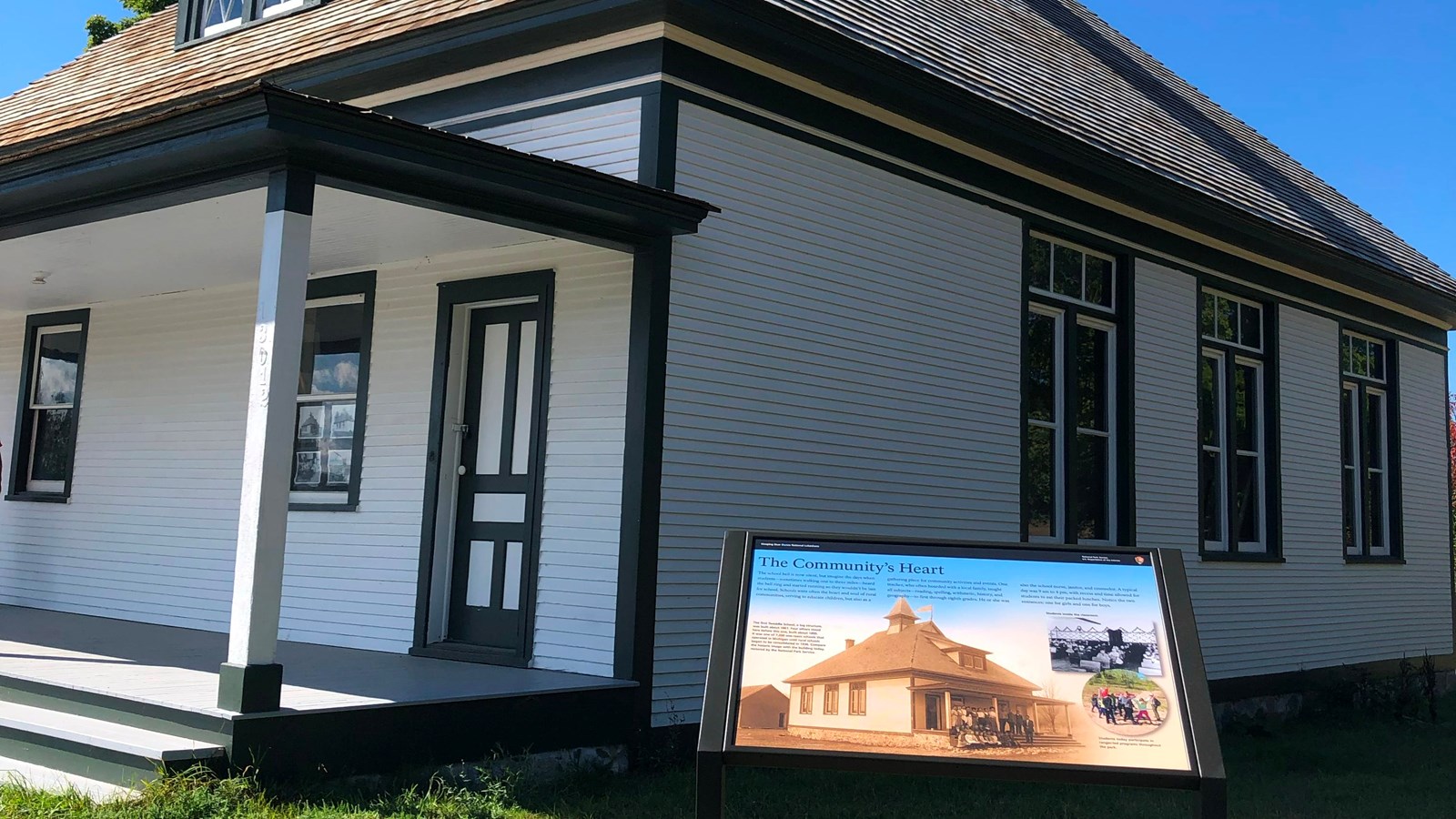 An informational sign stands on a grass lawn in front of a white two-story schoolhouse