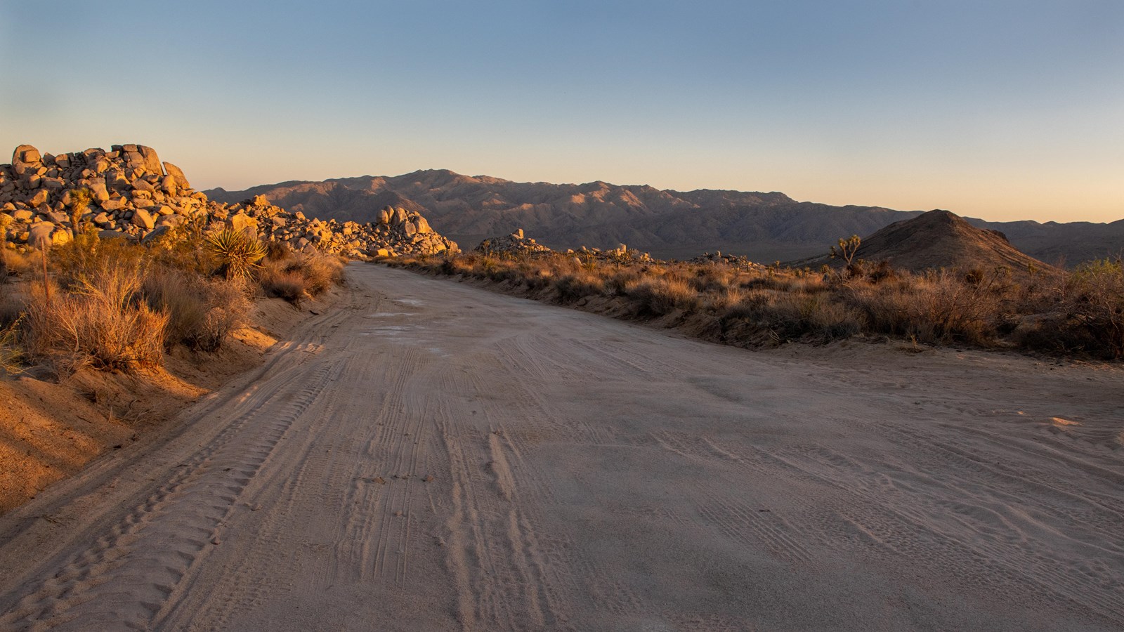 A dirt road leads towards rocks, a desert valley, and mountains.