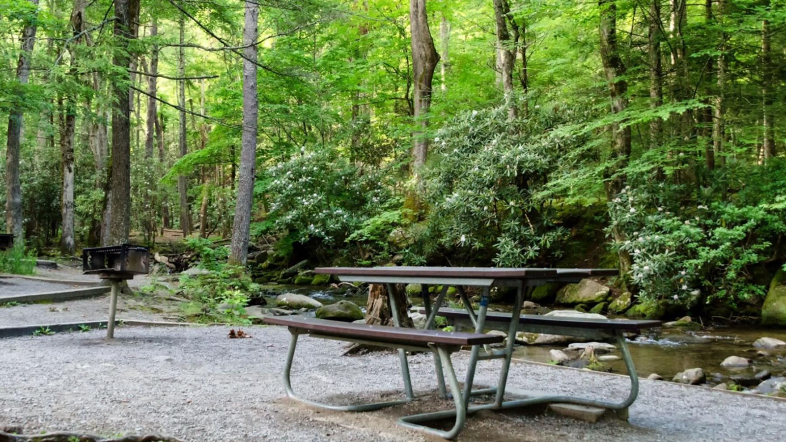 A wooden picnic table with a metal frame sitting on a gravel pad near a grill surrounded by trees.