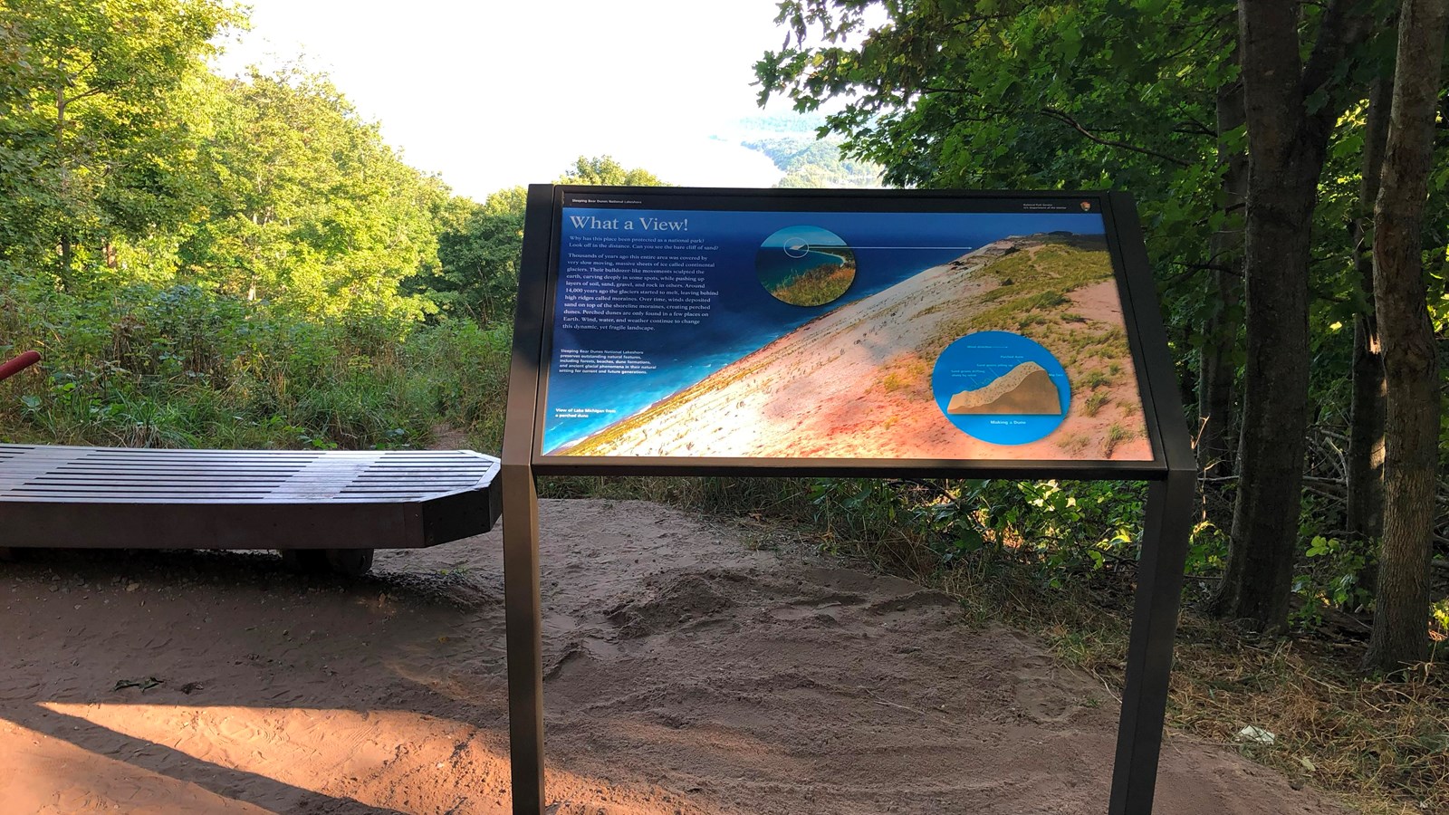 An informational sign stands to the side of a bench at a viewpoint along a forested trail