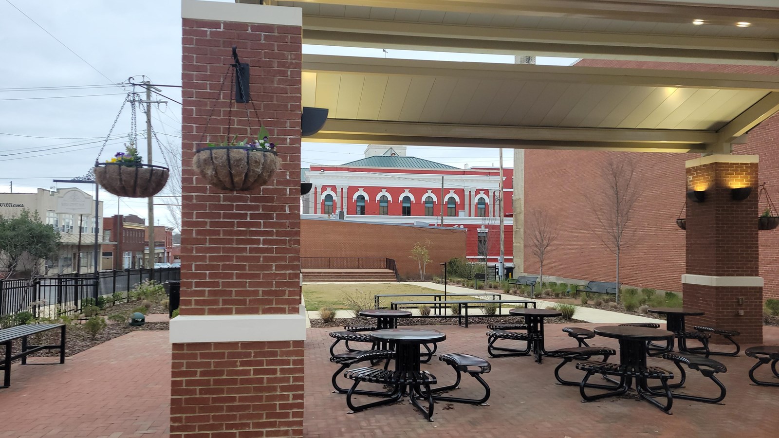 A brick and metal pavilion with picnic tables. Benches and Lanscaping surround the pavillion.