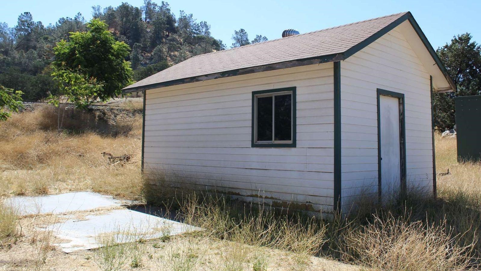 A small white shed with dark trim. A small concrete pad to the left of the building. Trees in back.