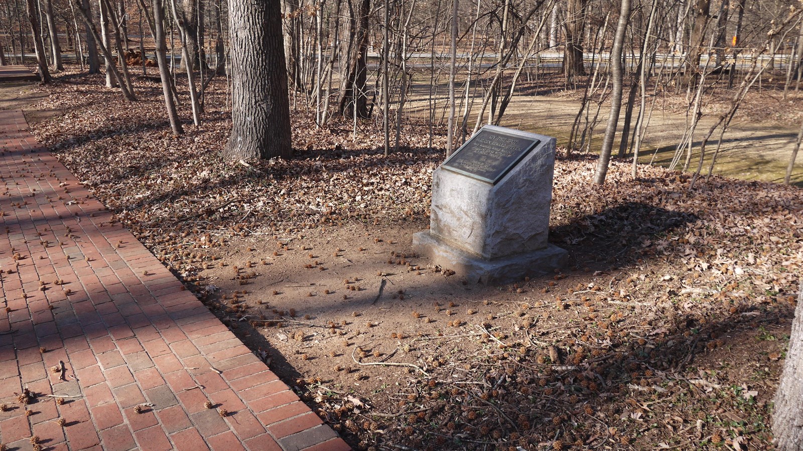 A short monument stands near a brick walkway, the New Garden trail in 5 yards behind it.