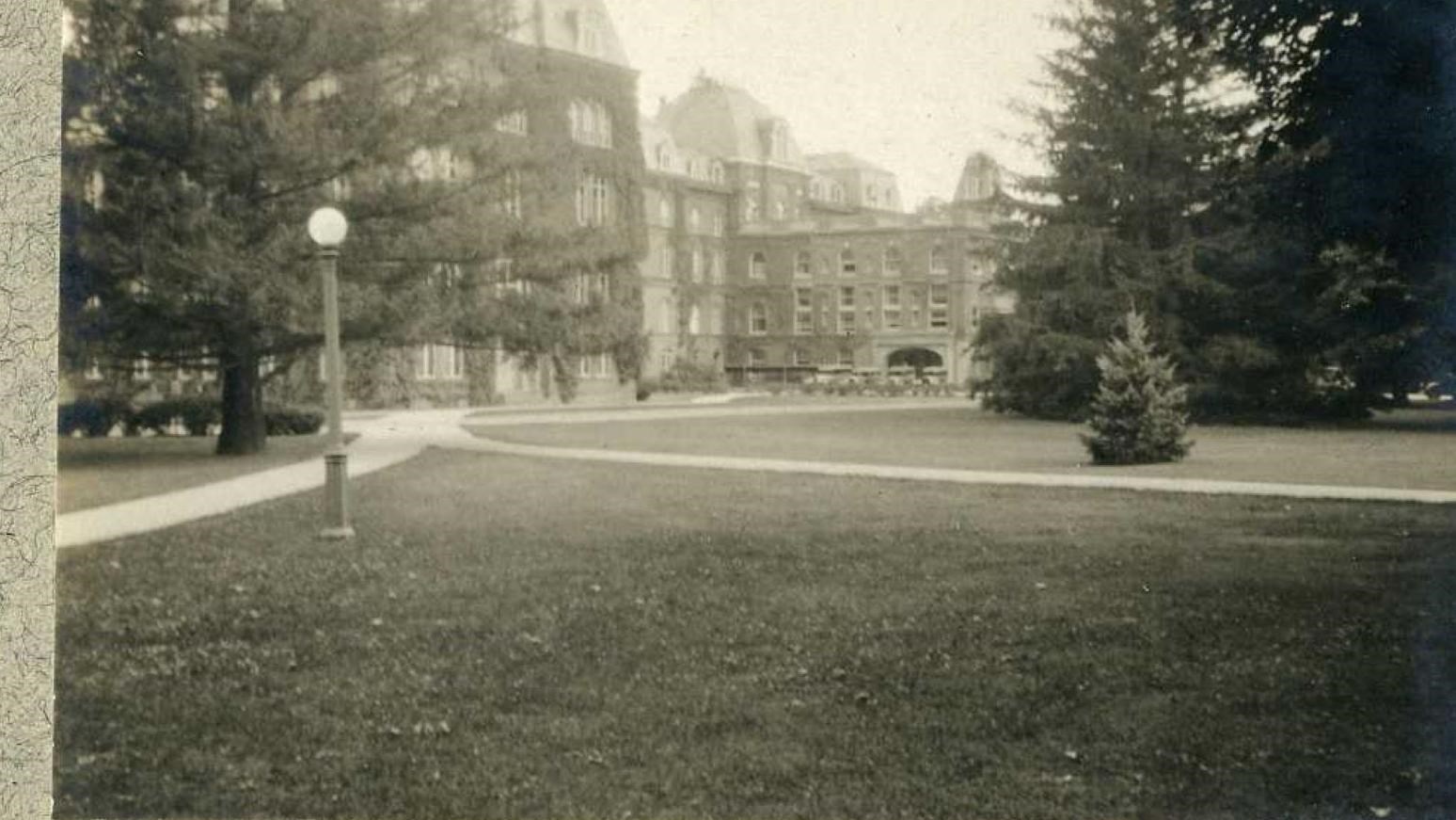 Black and white of flat grassy area with paths cutting across it, trees along parts, building behind