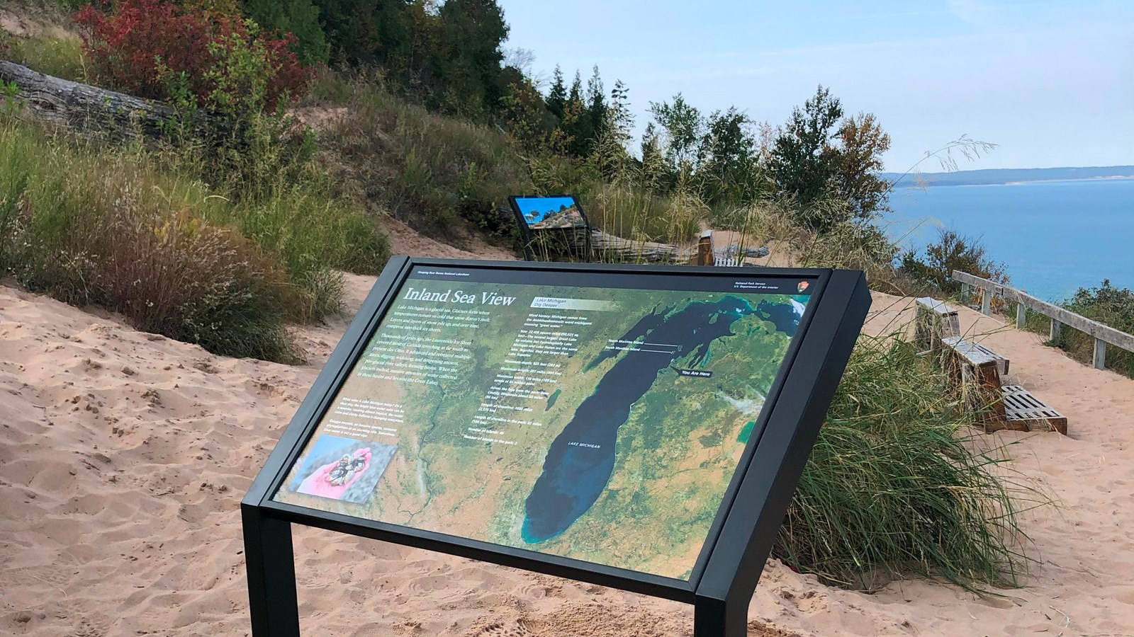 An informational sign stands along the sandy slope of a dune. Plants cover the dune behind it.