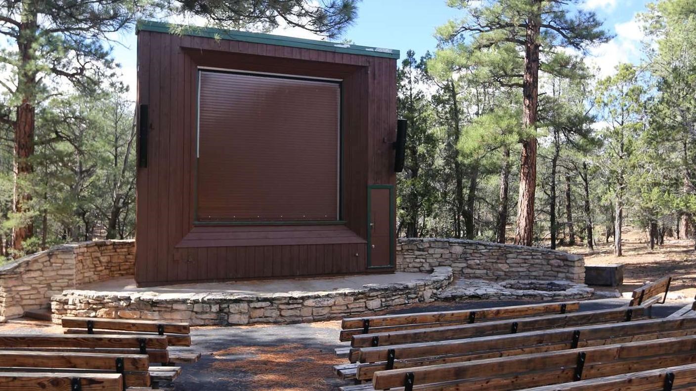 A large outdoor screen protected by a brown building sits in front of row after row of benches.