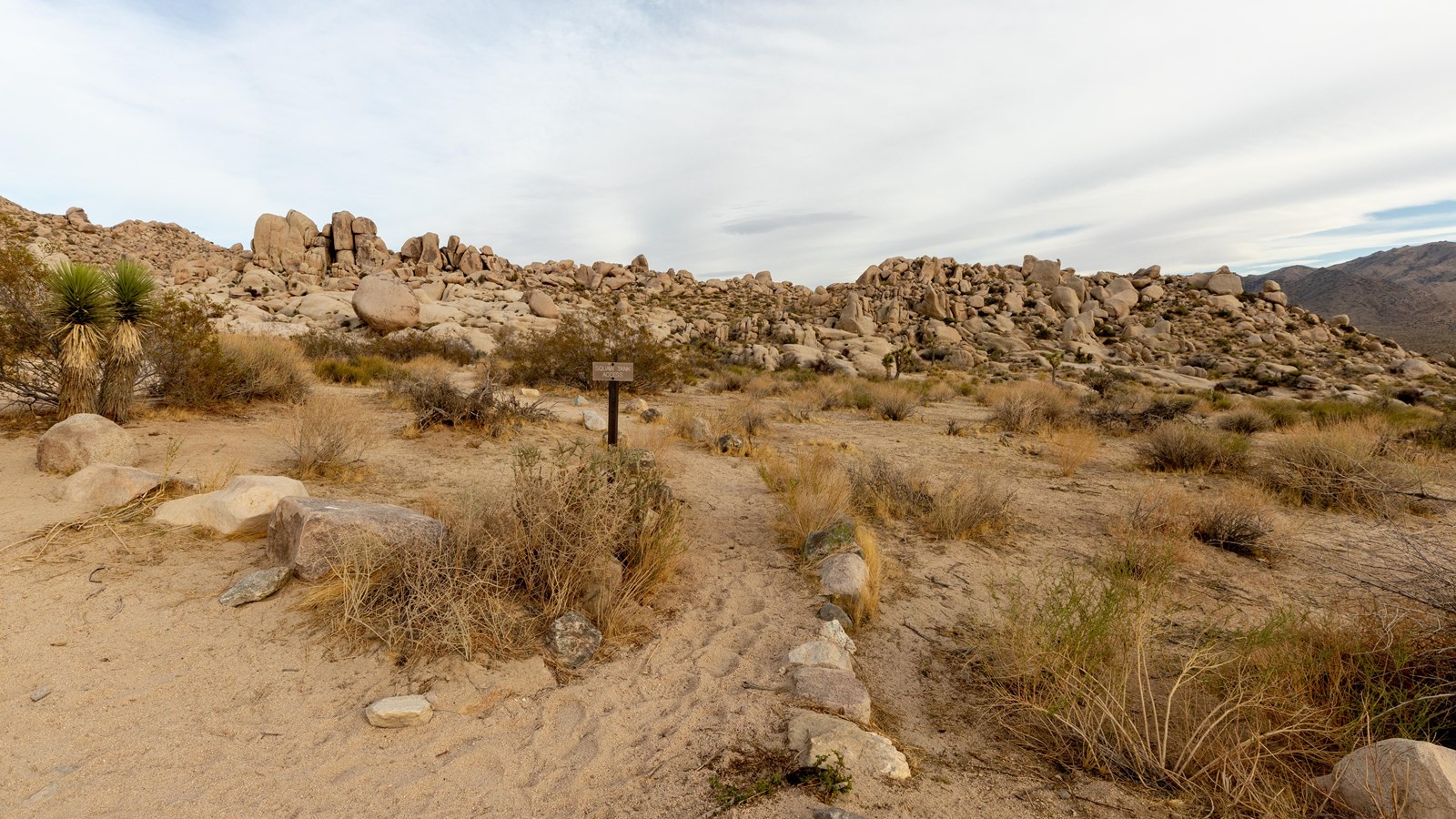 A trail sign next to a small, dirt trail, surrounded by rocks.