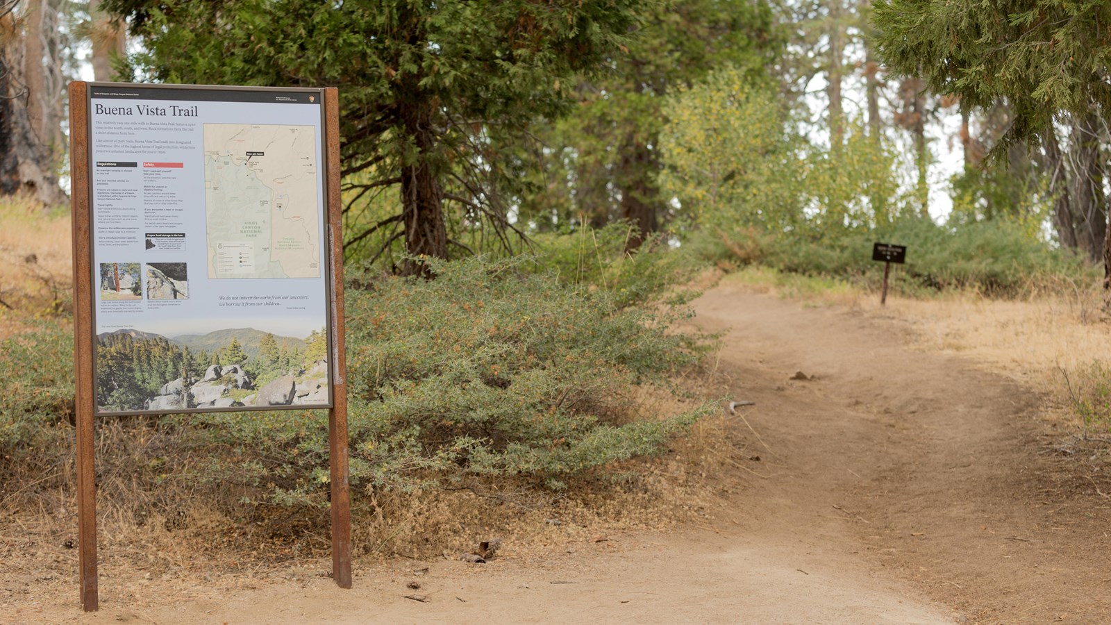 A metal framed sign stands to the side of a dirt trail. Behind the sign are bushes and trees.
