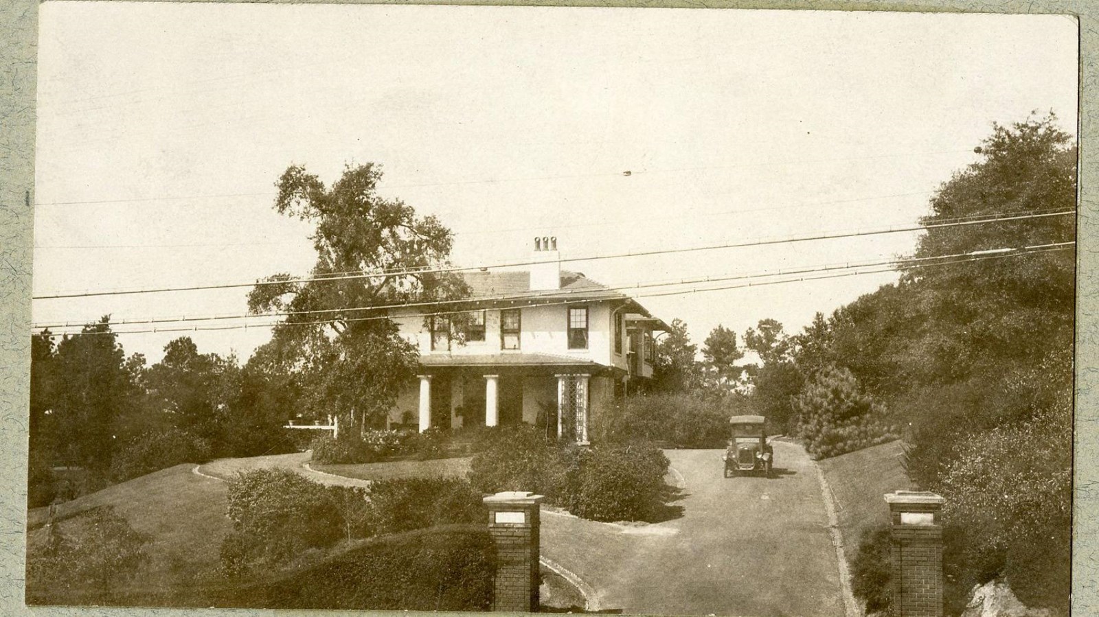 Black and white of home on top of grassy hill with trees and shrubs on hill, car in driveway