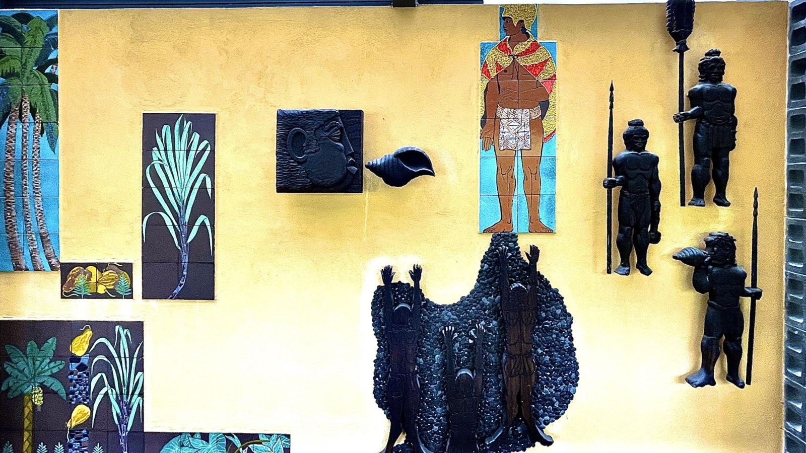 Carving and painting depicting men with spears around a chiefly man dressed in yellow feather cape.