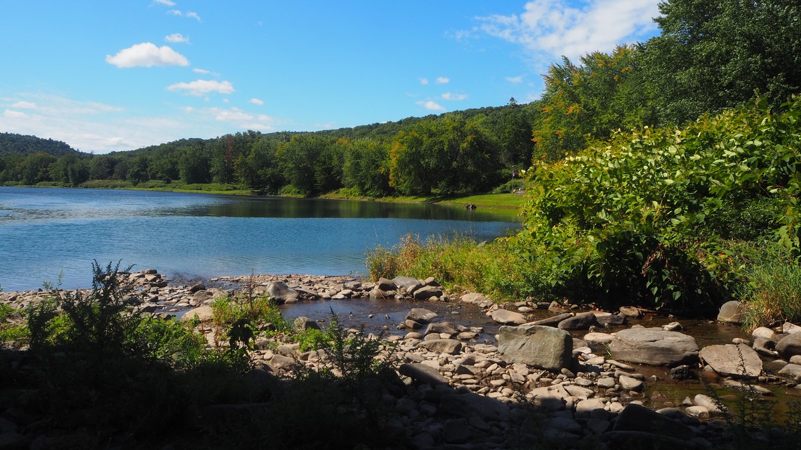 Blue river reflecting sky. Green grass and trees cover hills. In foreground, rocky stream.