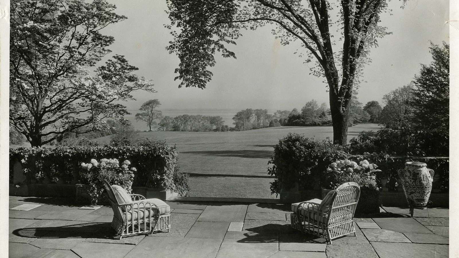 Black and white of two chairs on brick patio facing large flat open grassy area, trees on edge