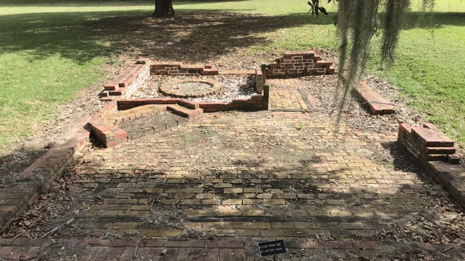 Rectangular brick foundation and floor with remnants of a fireplace and oven; grassy area surrounds.