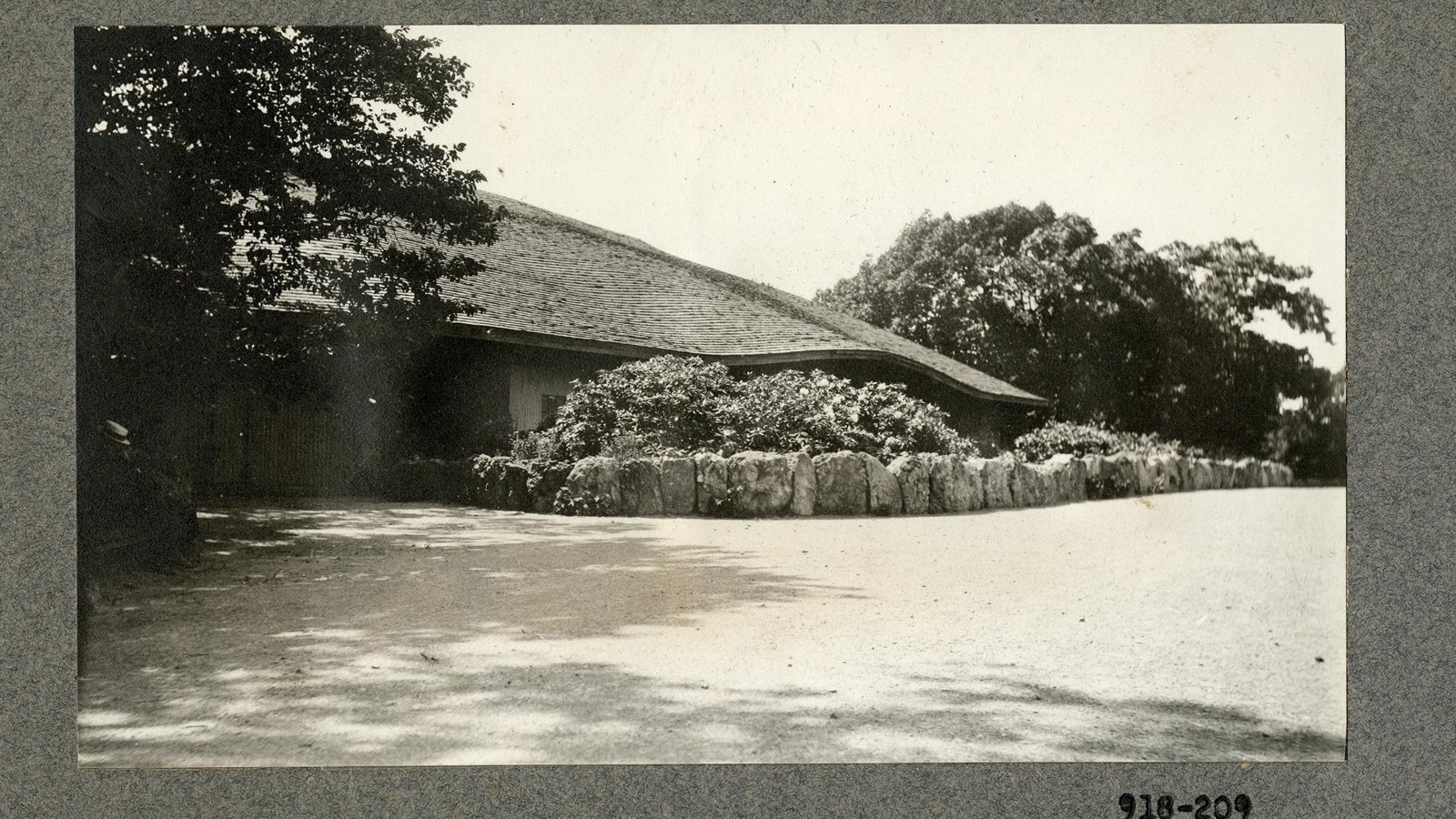 Black and white of low building with down slanting roof with rock wall in front and shrubs
