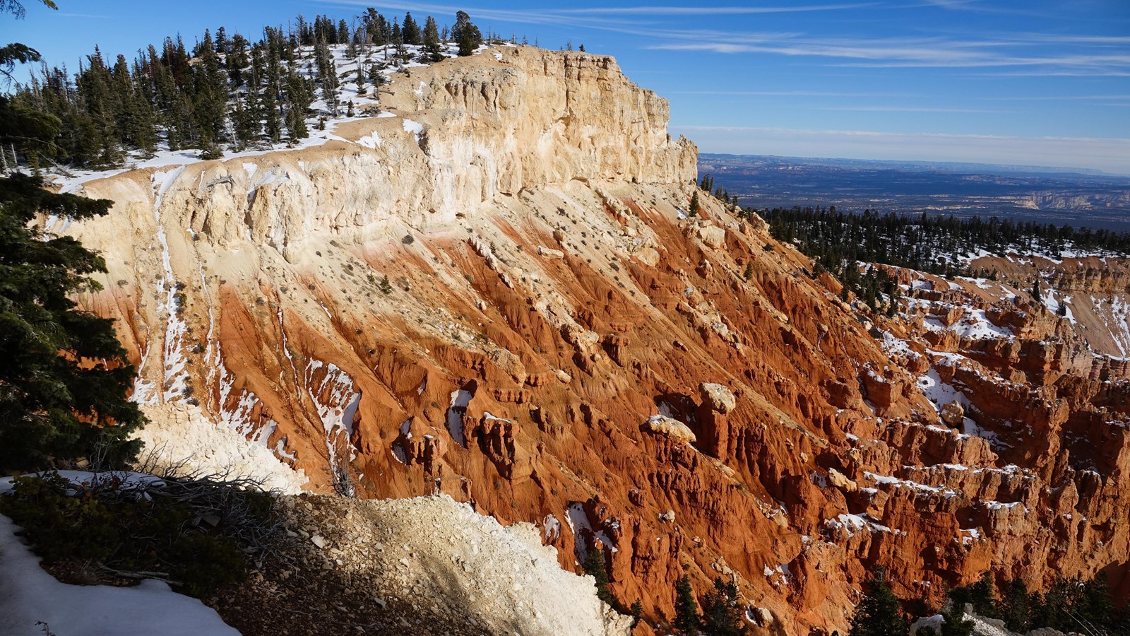 A brightly colored cliff of orange and white rock with a forested cliff above