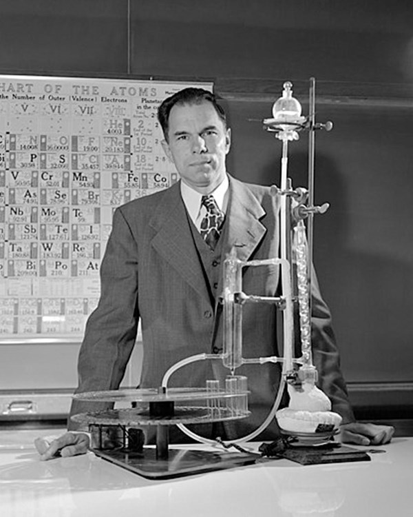 A man in a suit stands behind lab equipment. A period table chart is in the background.