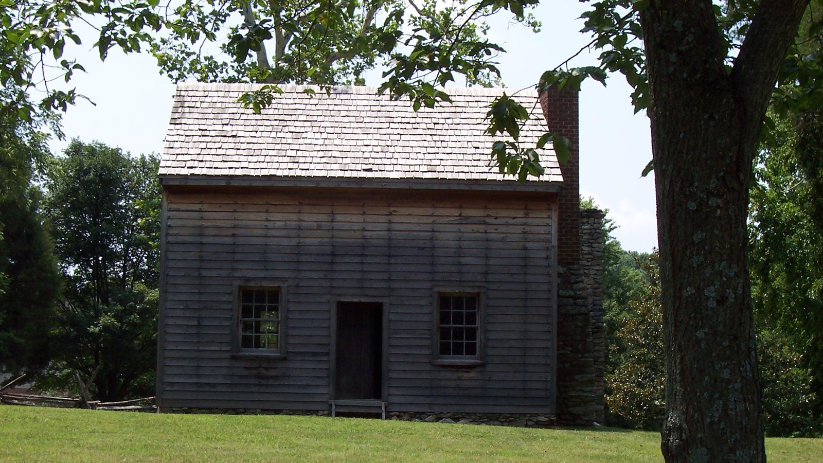 A two story colonial home with clap board siding stands in the open. Trees are visible behind it.