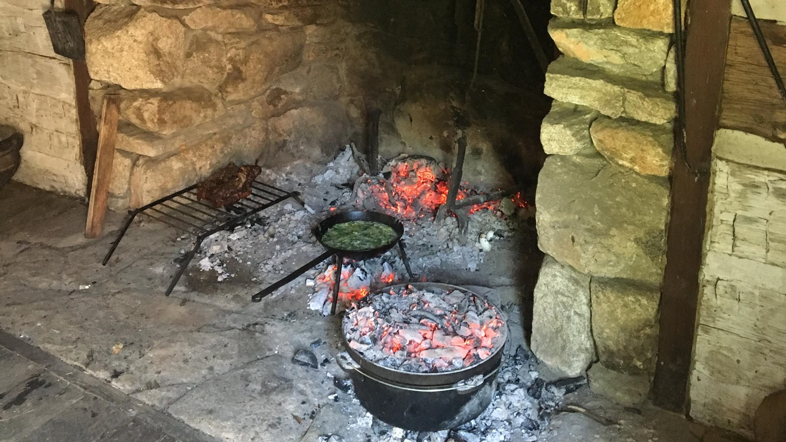 In a colonial hearth a fire crackles. Several pots and a Dutch oven sit and cook over the coals.