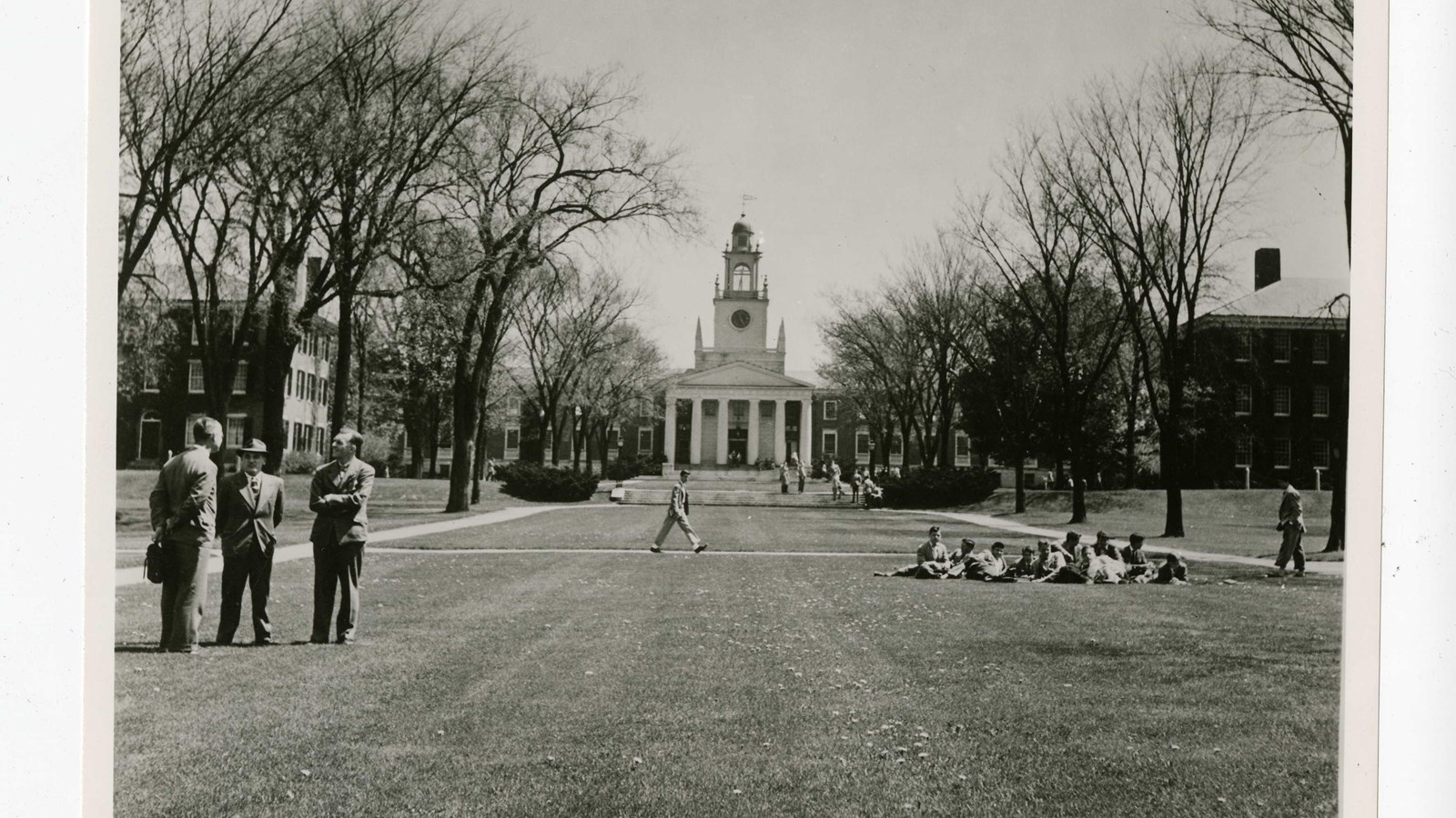 Black and white of flat grassy area with paths on both sides lined with trees, building at end 