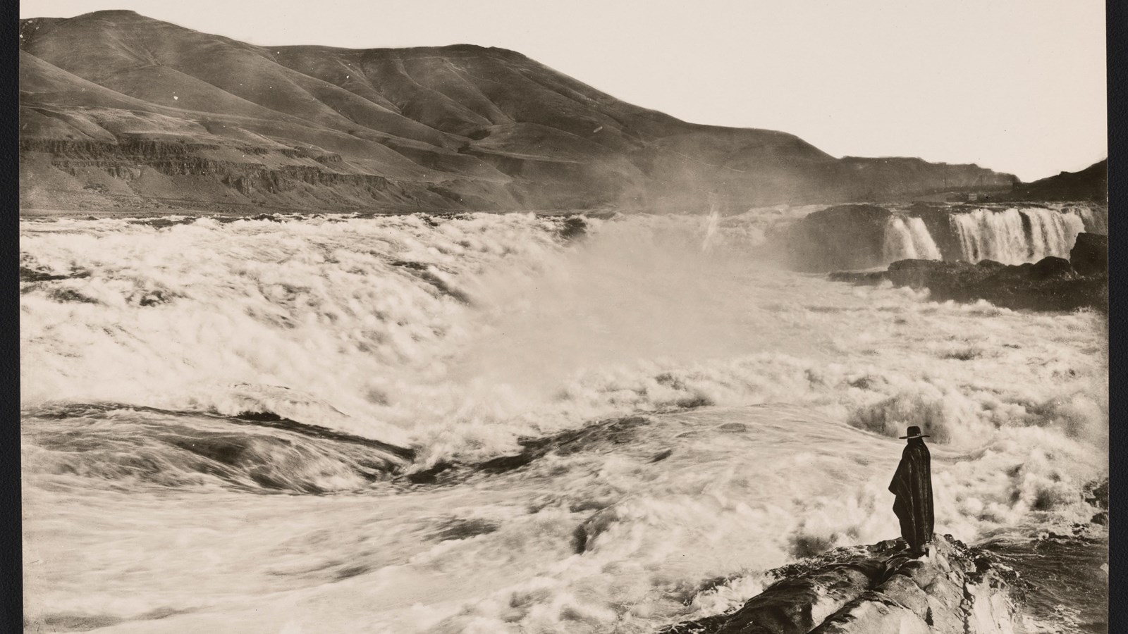 Waterfall churning water over rocks. Man stands in foreground.
