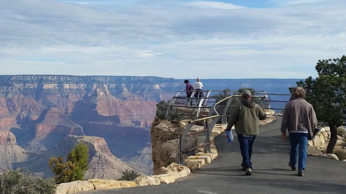 Two men walk down a paved path lined by rough hewn white rocks towards a fenced in viewpoint.