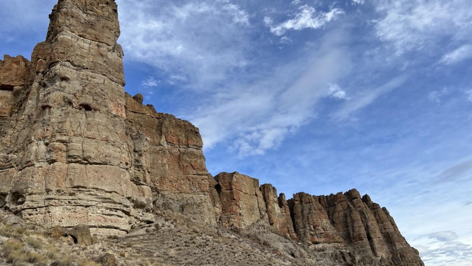 Tan cliff formations with a grassy slope below dotted with fallen boulders.