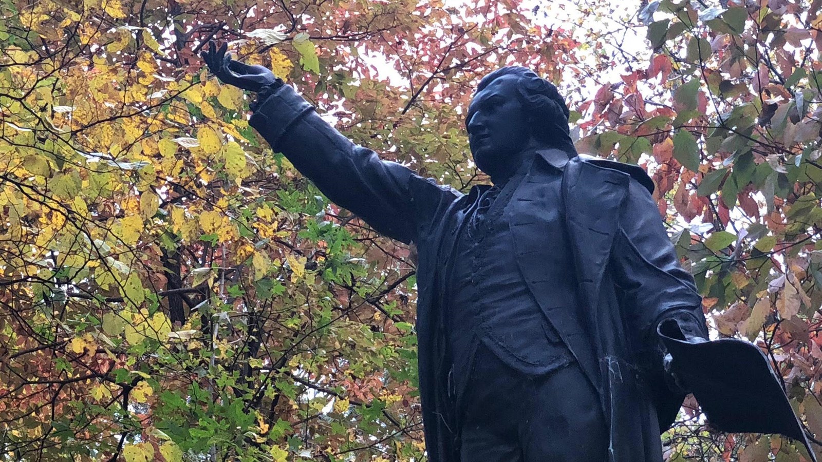 A bronze statue of William Hooper reaches to the sky, surrounded by fall foliage.