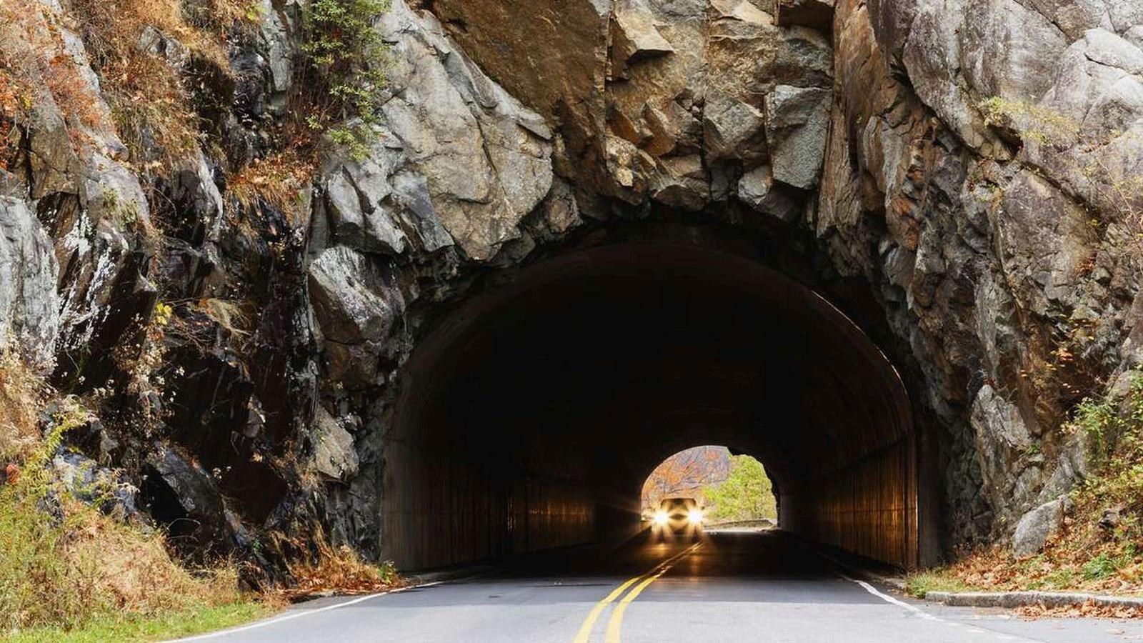 A tunnel cut into the mountain with trees on top of the rock tunnel exterior and a car\'s headlights 