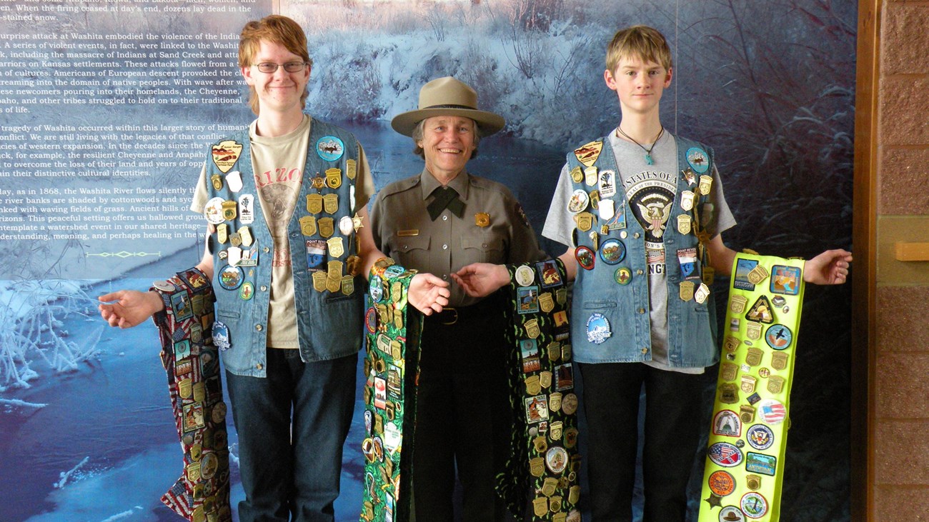 A ranger standing between two junior rangers with vests and banners covered in junior ranger badges.