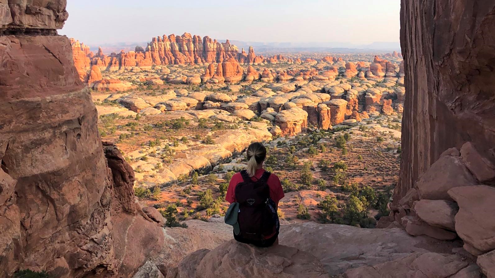 A hiker sits atop a sandstone rock looking out across a vast view of sandstone spires. 