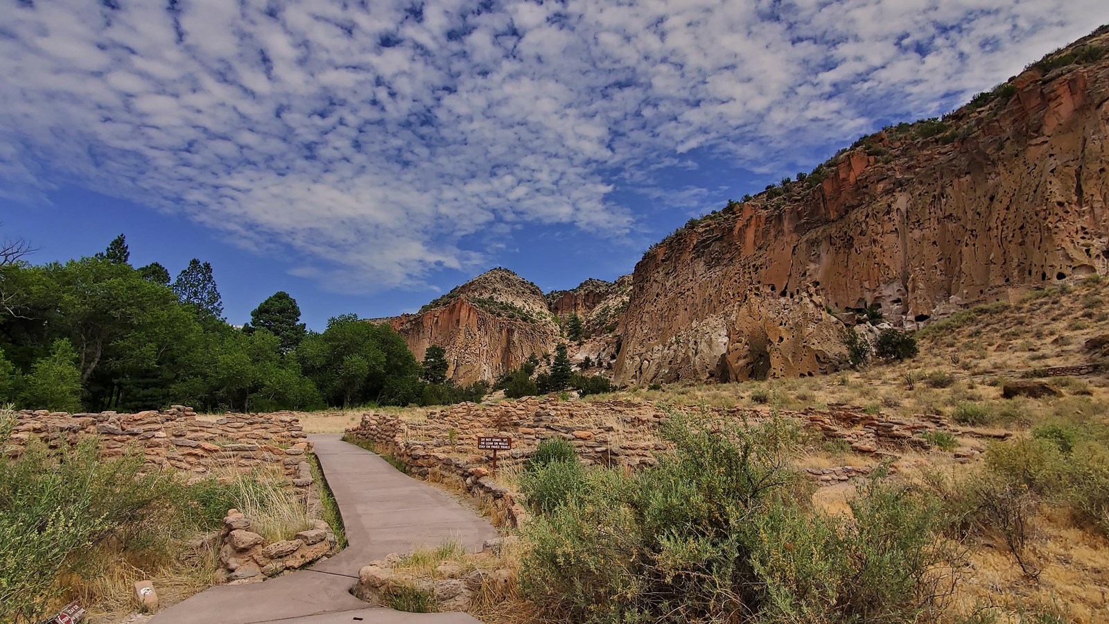 a paved trail enter a stone village with cliffs around and a cloudy blue sky