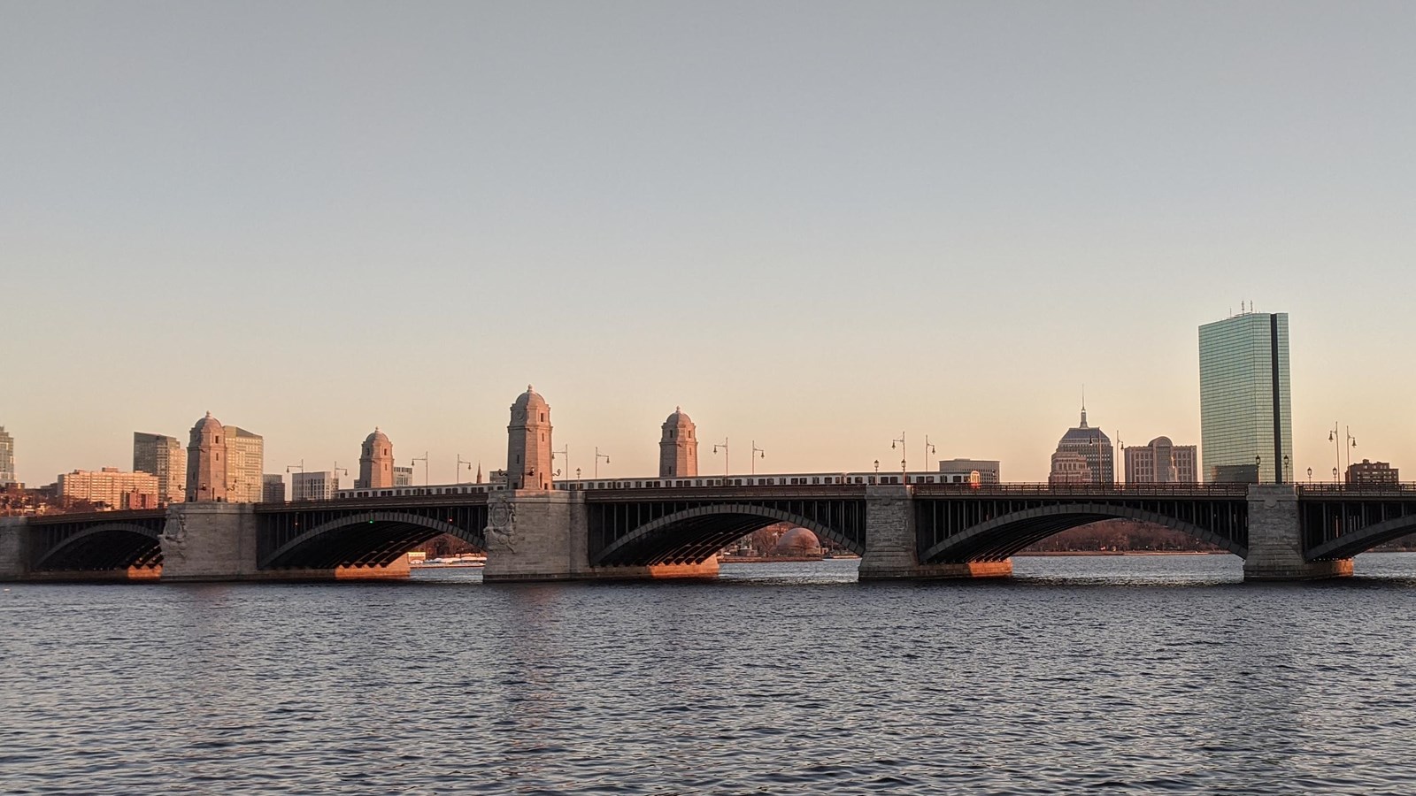 Bridge over river with stone supports and metal arches. Boston skyline in background.