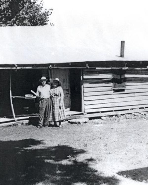 Black and white image of a man and woman standing outside of a log cabin