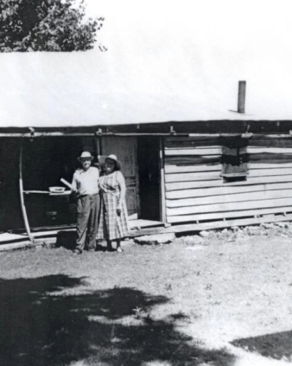 Black and white image of a man and woman standing outside of a log cabin