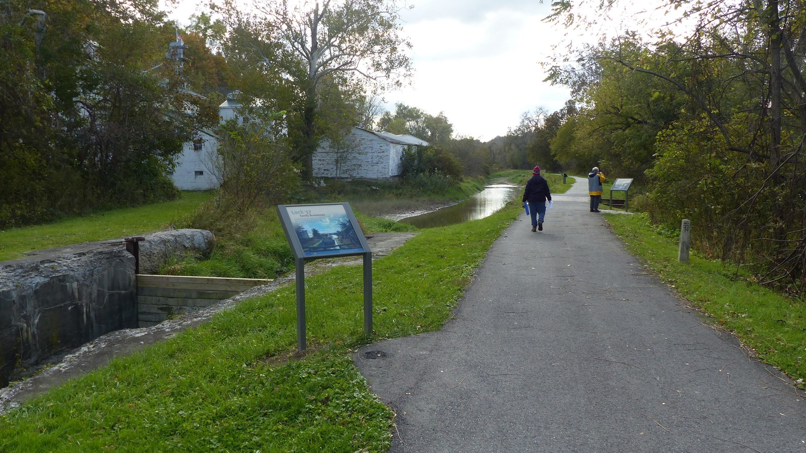 Two people walk a paved trail, pausing to read graphic panels. On the left is a water-filled canal w