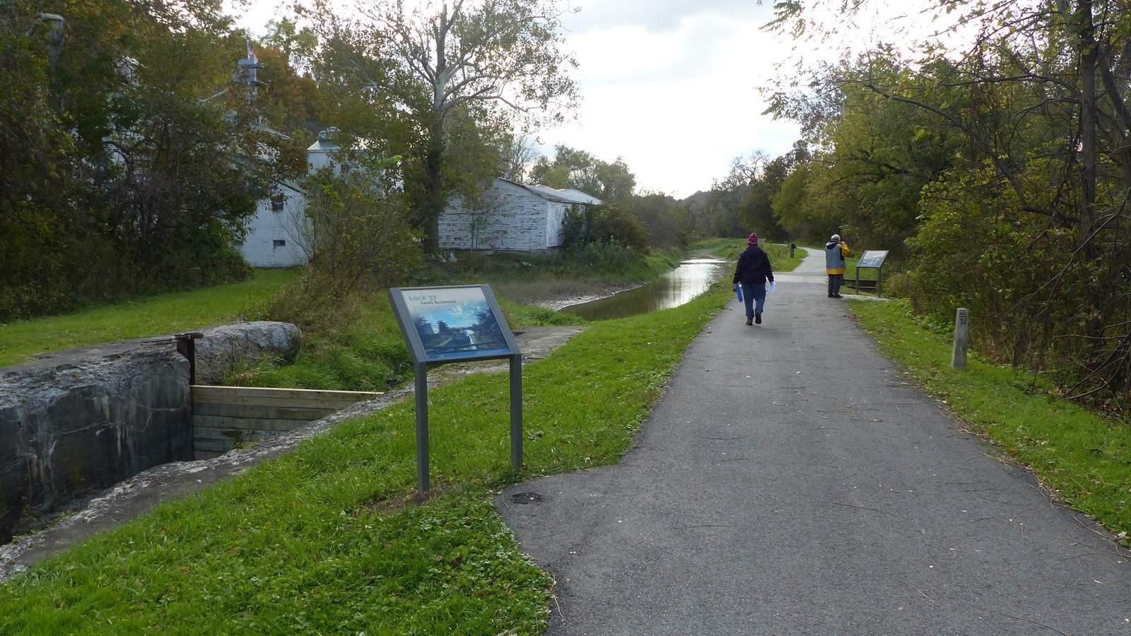 Two people walk a paved trail, pausing to read graphic panels. On the left is a water-filled canal w