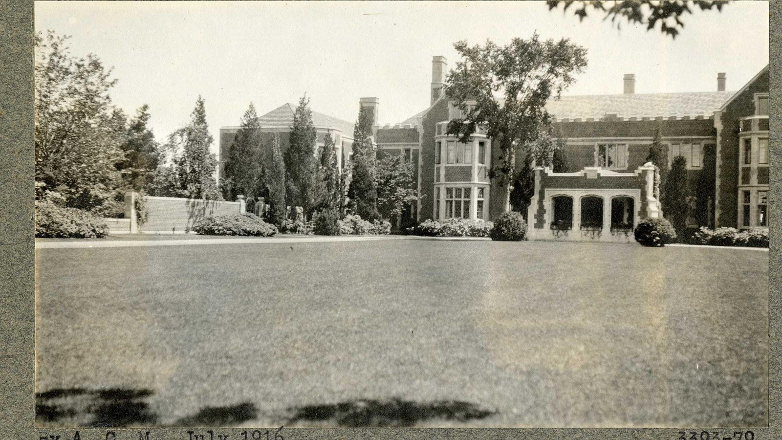 Black and white of flat grassy area in front of large brick house with trees and shrubs around it