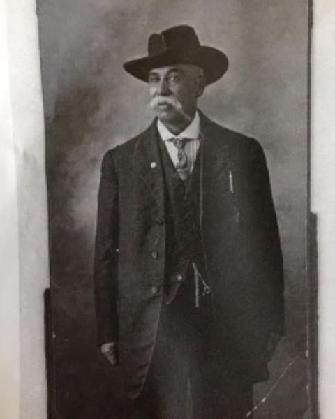 Man wears brimmed hat and three piece suit.  He had a white mustache and tie.