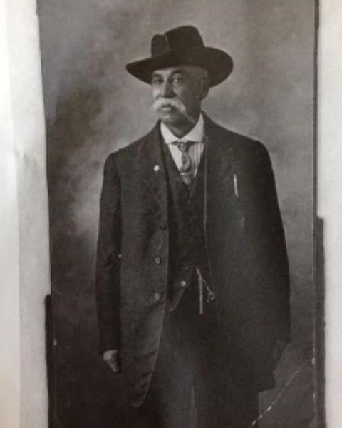 Man wears brimmed hat and three piece suit. He had a white mustache and tie.