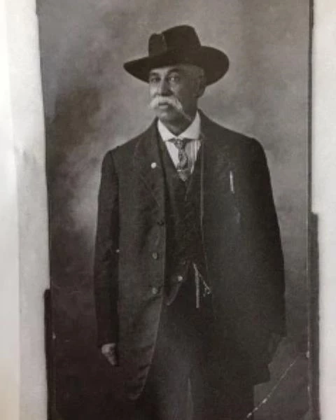 Man wears brimmed hat and three piece suit.  He had a white mustache and tie.