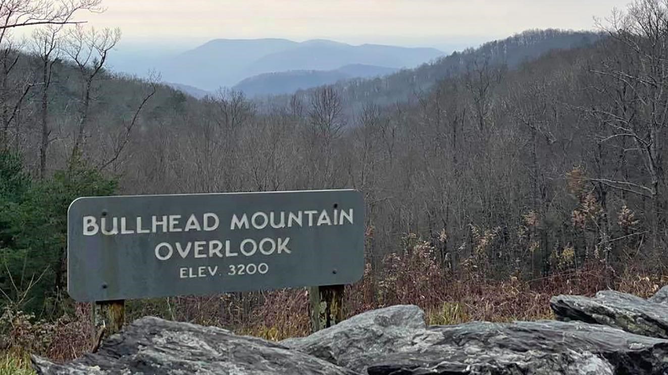 A rock wall and sign stand in front of a bare, winter forest, with mountains in the distance