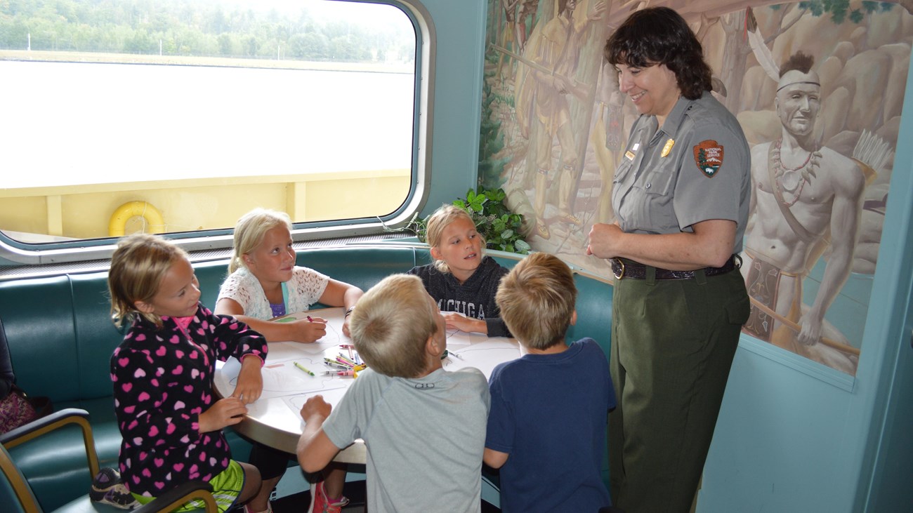 Children sit around a table working on Junior Ranger booklets while a Park Ranger stands nearby.