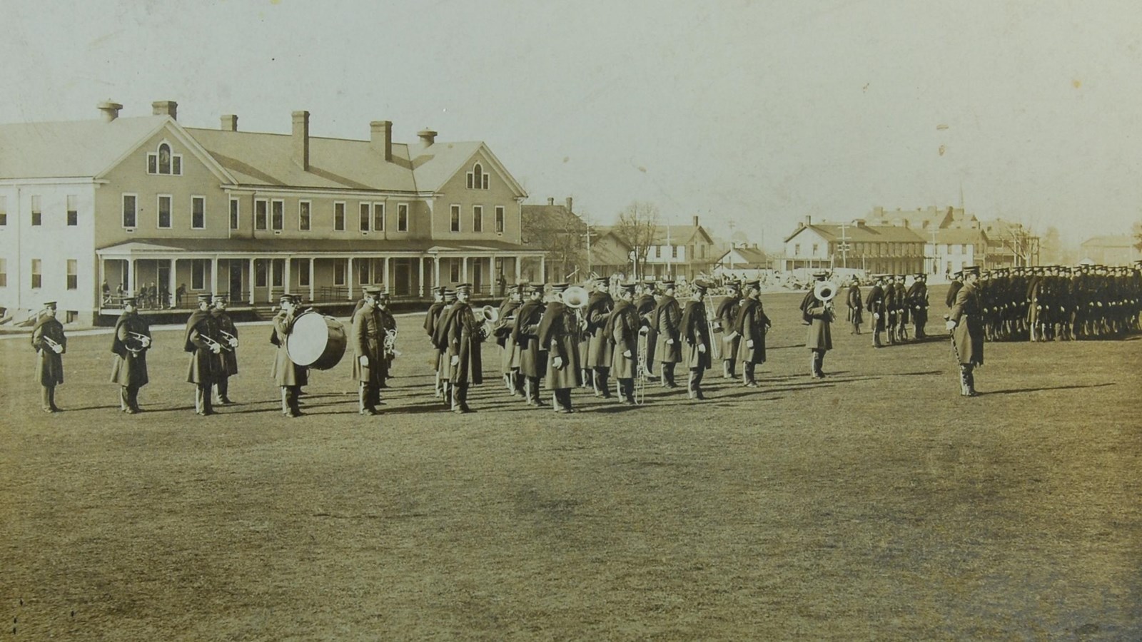 A black and white photo shows a military band standing on a field with barracks buildings behind.
